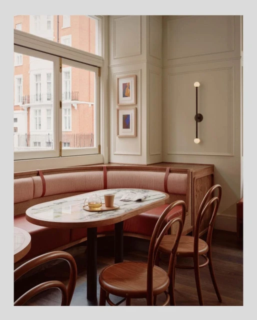 Interior corner of a restaurant with a marble-topped oval table set for two, surrounded by curved wooden chairs and bench seating.