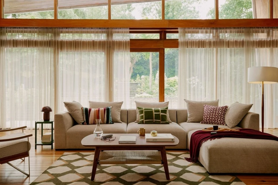 Living room interior featuring a tan sectional sofa with throw pillows, wooden coffee table, floor lamp, area rug and sheer curtains.