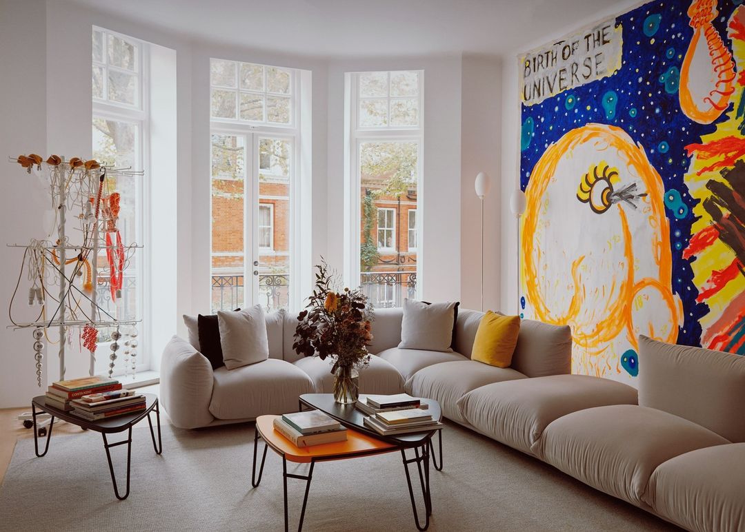 Living room features beige sectional sofa, abstract wall art, coffee tables with books, and white-framed windows showcasing an outdoor view.