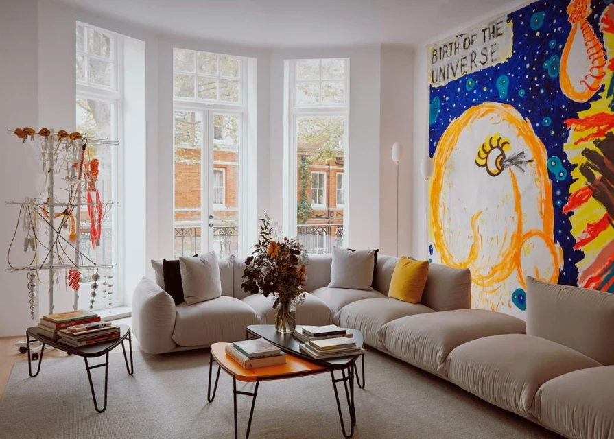 Living room features beige sectional sofa, abstract wall art, coffee tables with books, and white-framed windows showcasing an outdoor view.