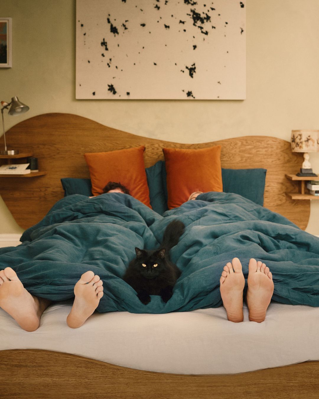 Two people under a blanket with a black cat, only their feet are visible at the end of the bed in a neutral-toned room.