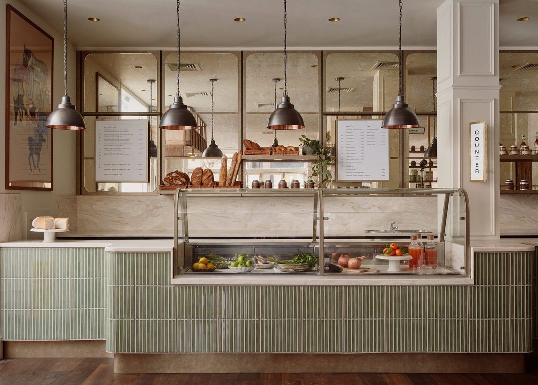 Indoor view of bakery display case and countertop with pastries, bread, fruit, and beverages beneath hanging pendant lights.