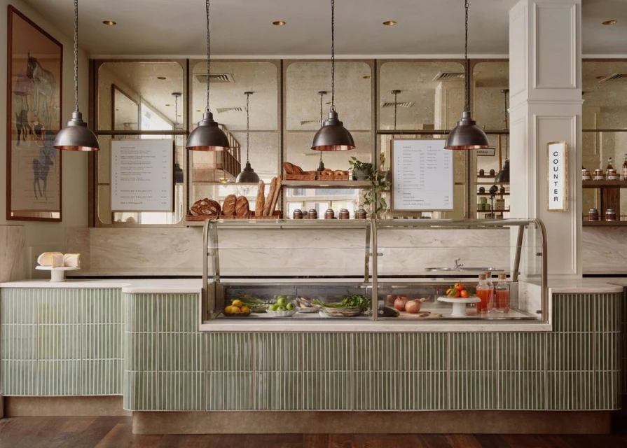 Indoor view of bakery display case and countertop with pastries, bread, fruit, and beverages beneath hanging pendant lights.