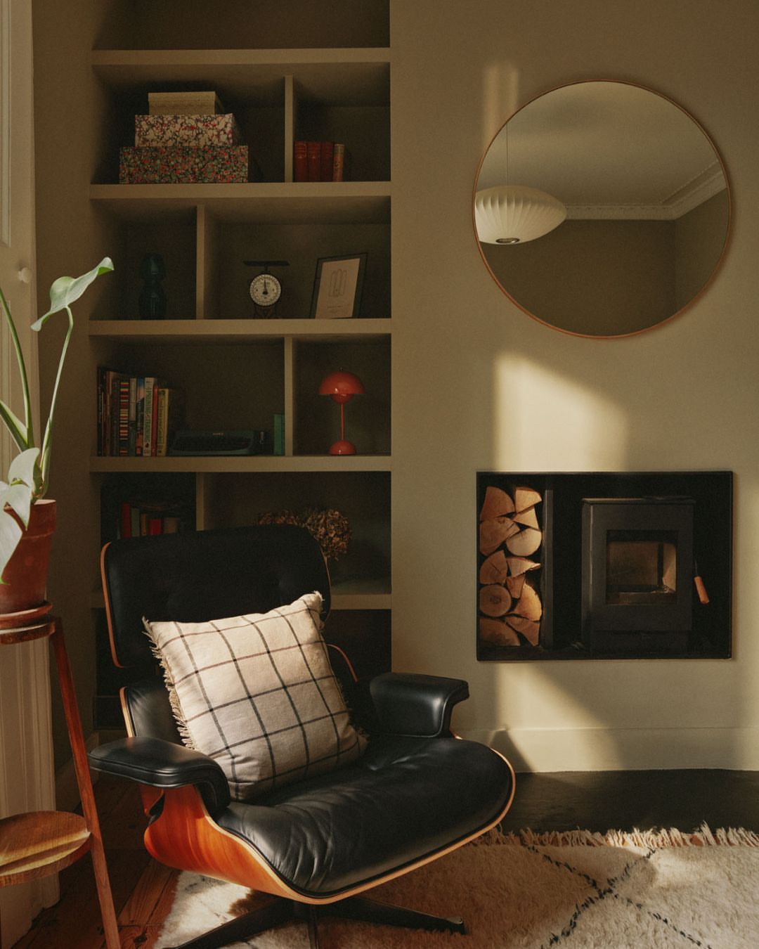 Living room interior with black lounge chair, wood fireplace, bookshelves, round mirror, indoor plant, soft natural lighting.