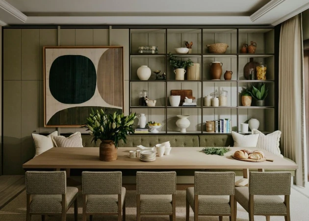 Interior of a dining room featuring a large wooden table with chairs, a vase of white lilies, and built-in shelving filled with dishware.