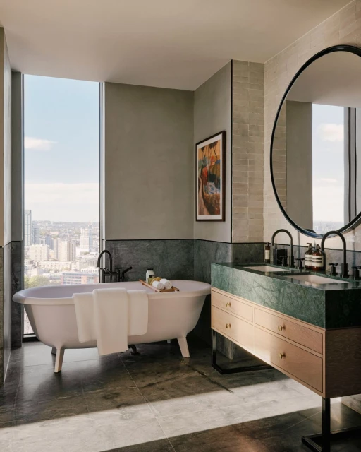 Bathroom interior featuring a white freestanding bathtub, marble vanity, round mirror, and a large window overlooking a cityscape.