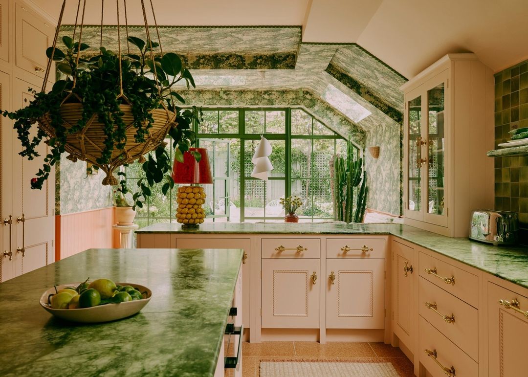 Cozy kitchen interior with green marble countertops, pink cabinetry, hanging plant, lamp, large window, and patterned wallpaper.