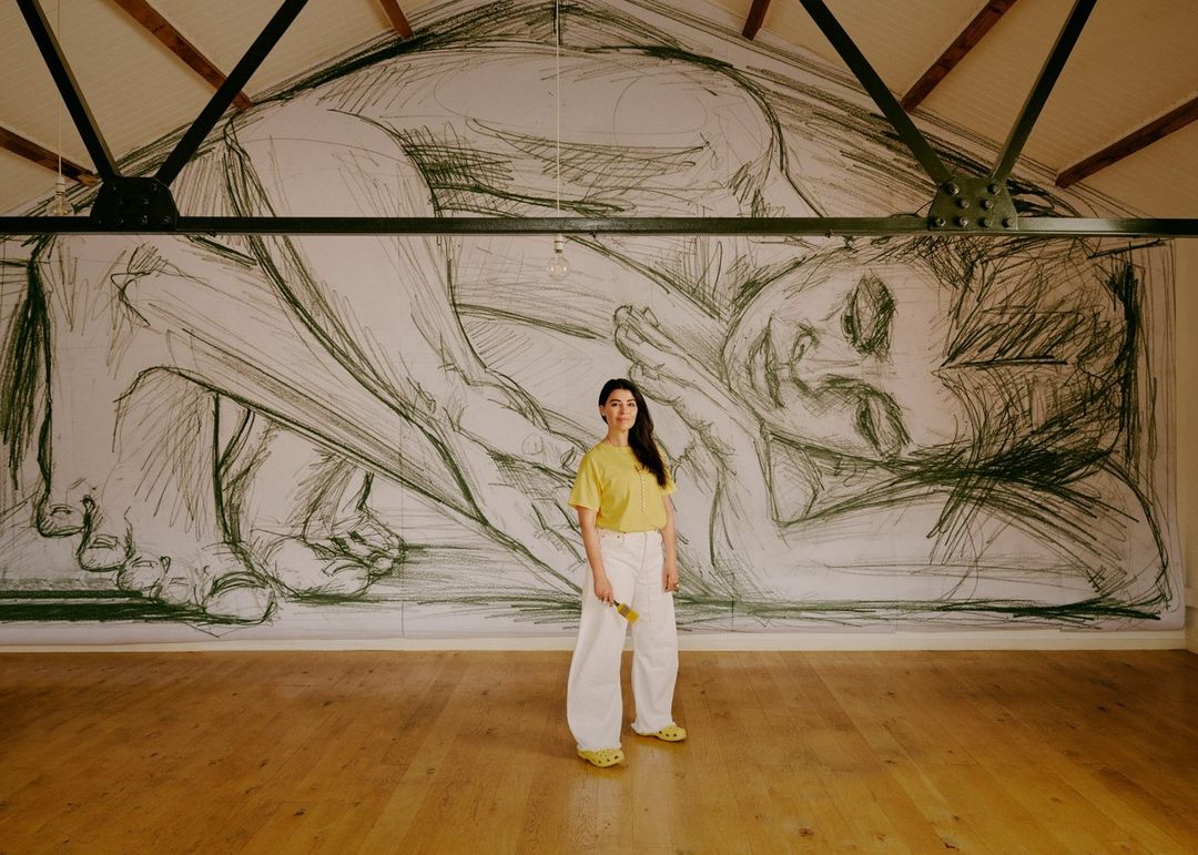 Woman poses in front of large sketch of a reclining figure in a room with a wooden floor and exposed ceiling beams.