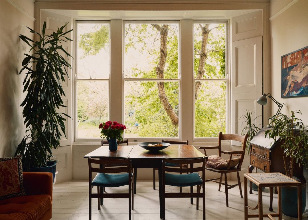 Brightly lit interior with a dining table and chairs, windows overlooking a lush garden, sofa, desk, and plants filling the space.