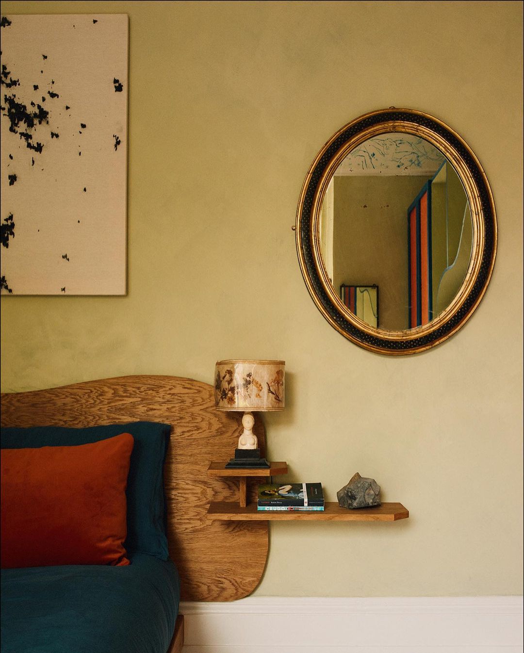 Bedroom interior features a bed with a wooden headboard, blue and orange pillows, a round mirror, and an abstract art piece on the wall.