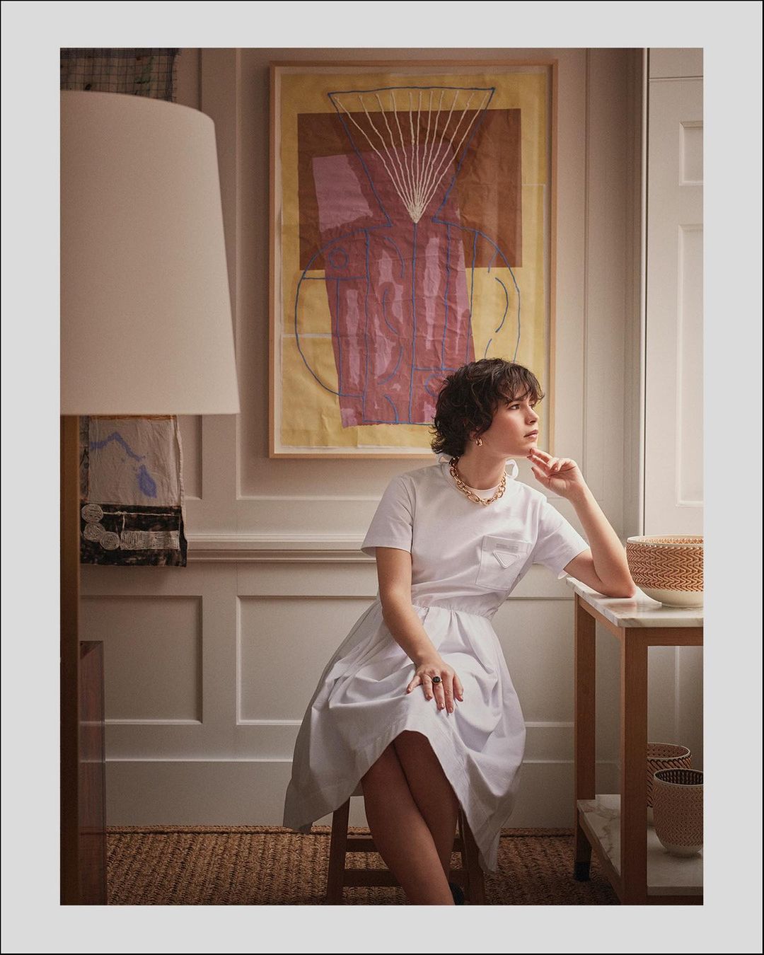 Woman in a white dress sits by a marble table in a sunlit room with neutral tones, complemented by woven baskets and abstract art.