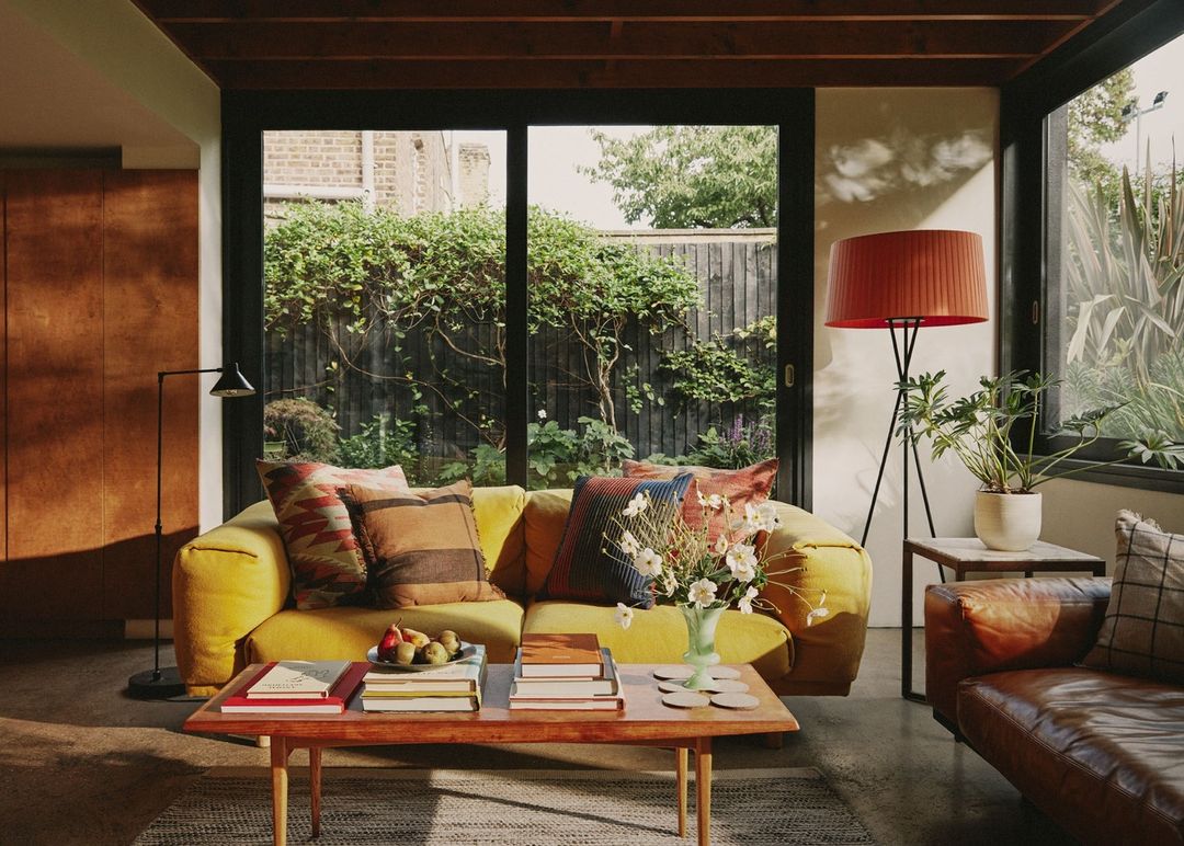 Cozy living room interior with a yellow sofa, patterned pillows, books on a coffee table, and a view to a green backyard.