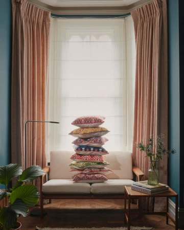 Living room interior featuring a modern sofa, a tall stack of decorative pillows, a side table, and stylish curtains. Natural light.