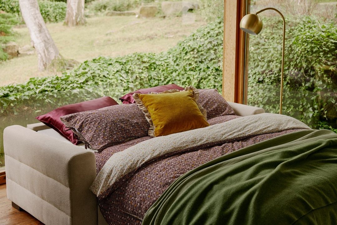 A cream-colored sofa bed draped with patterned bedding, a green blanket, and a mustard yellow accent pillow, positioned in a room with a large window overlooking lush greenery.