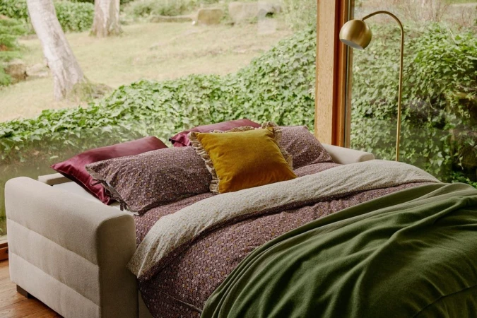 A cream-colored sofa bed draped with patterned bedding, a green blanket, and a mustard yellow accent pillow, positioned in a room with a large window overlooking lush greenery.