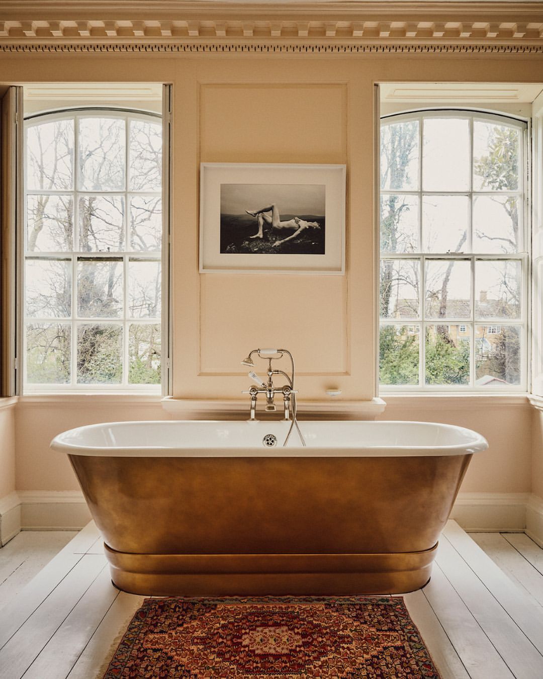 Freestanding bathtub in a beige bathroom with white trim and two large windows, next to a red patterned rug on a white floor.