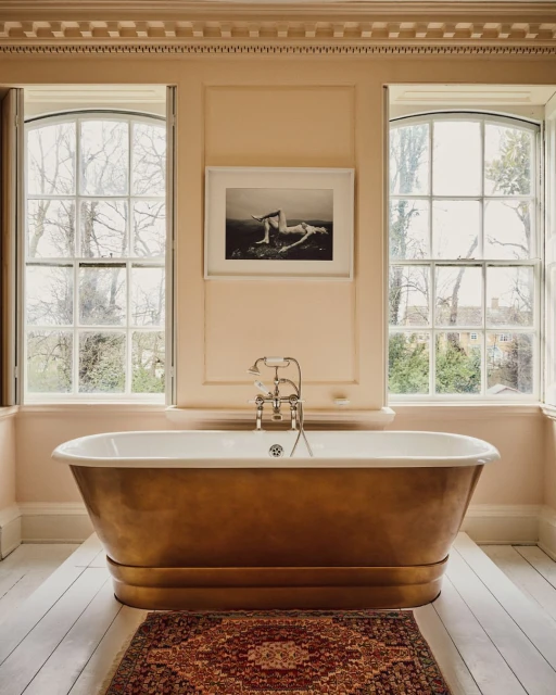 Freestanding bathtub in a beige bathroom with white trim and two large windows, next to a red patterned rug on a white floor.