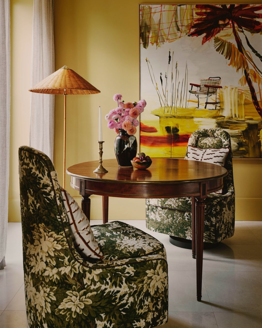 Interior design featuring a round wooden table, floral-patterned chairs, and a painting with a tropical theme, complemented by a floor lamp.