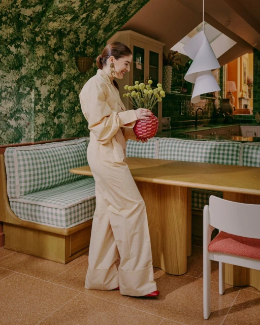 Woman in a light yellow jumpsuit holds a vase of yellow flowers indoors, next to a booth with green and white checkered cushions.