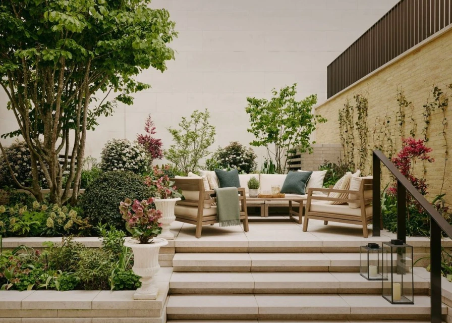 Outdoor patio area features a neutral cushioned sofa and chairs, greenery, and a stone staircase in daylight, creating a serene space.