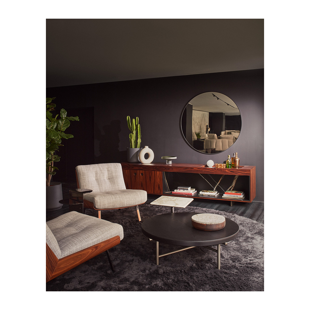 Living room interior with armchairs, round coffee table, and a wooden cabinet with a round mirror in the background.