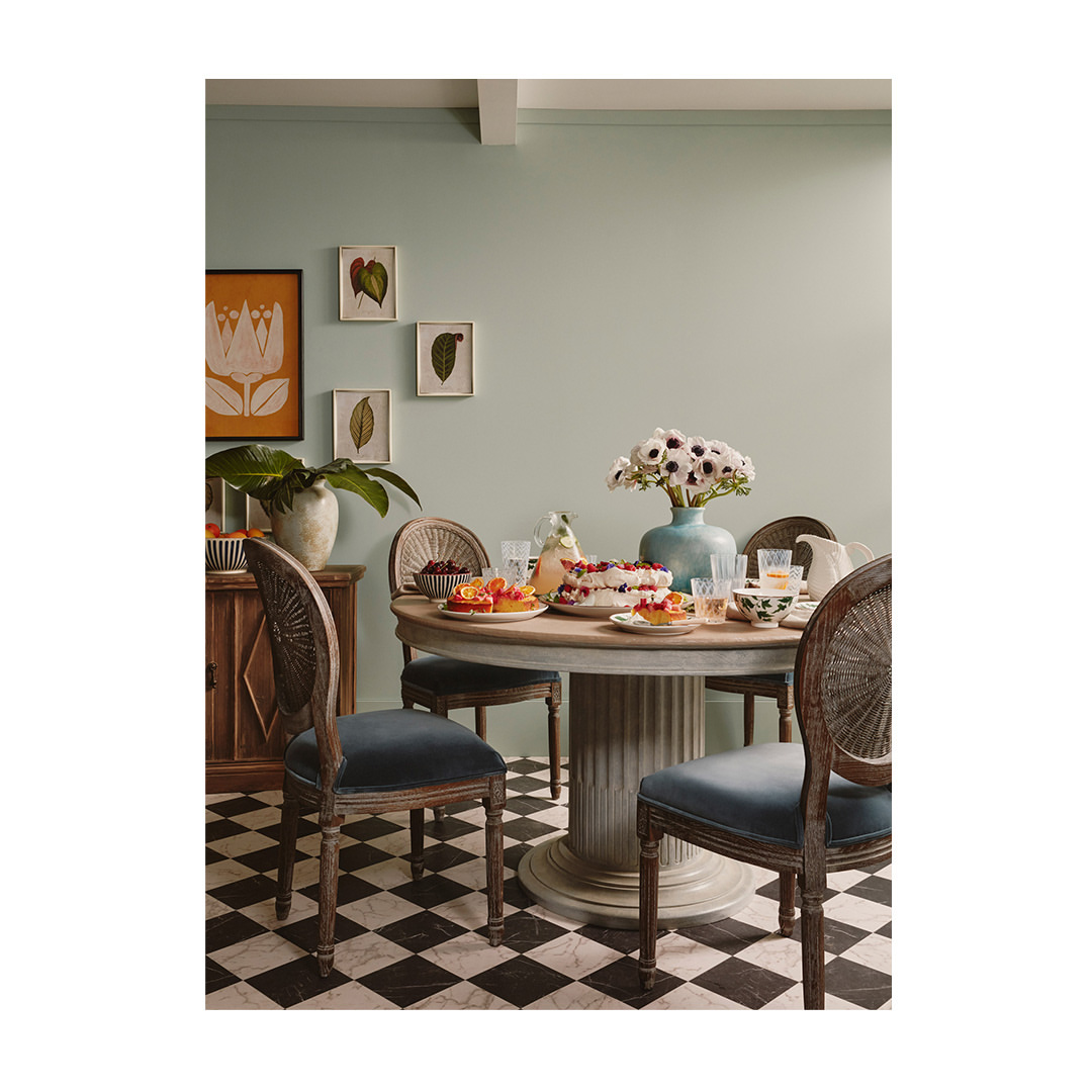Round table set with a fruit dessert, plates of fruit, glasses of water, and a flower arrangement with chairs in a room.