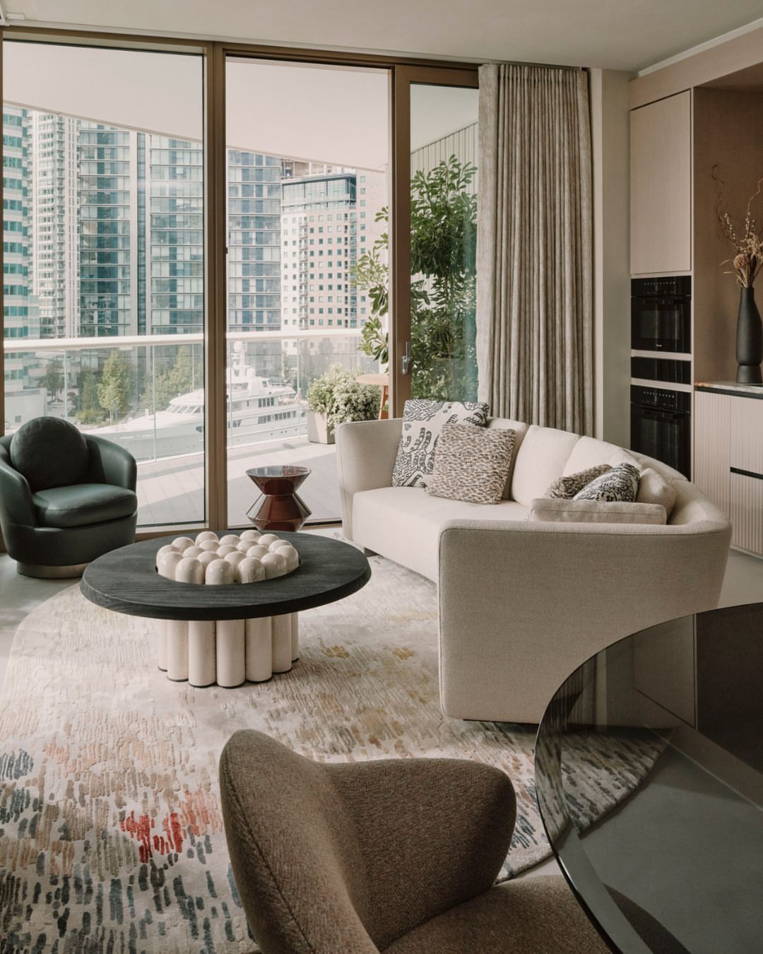 Modern apartment interior featuring a neutral curved sofa, round coffee table, cityscape view through large windows, and stylish decor.
