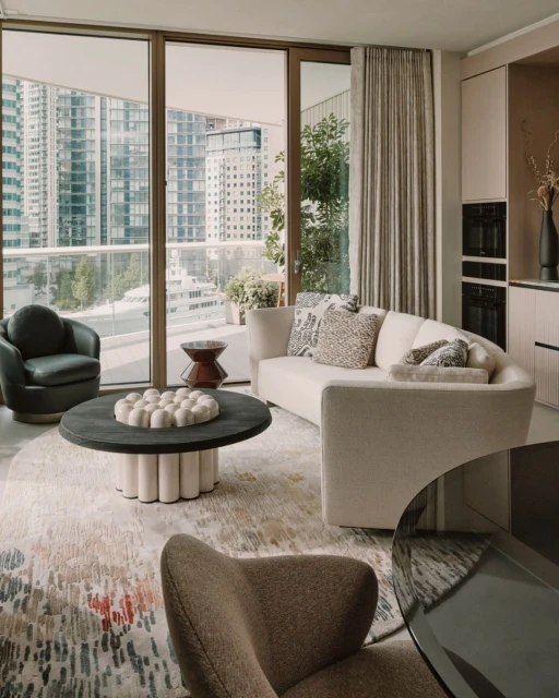 Modern apartment interior featuring a neutral curved sofa, round coffee table, cityscape view through large windows, and stylish decor.