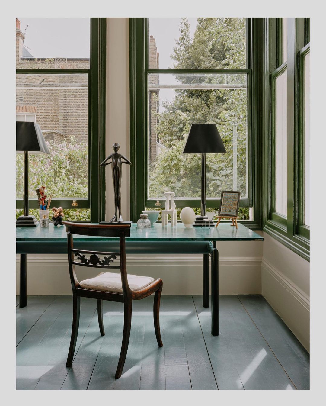 Interior view of a desk with lamps, a sculpture, and a chair by a window, showcasing a blend of classic and modern design elements.
