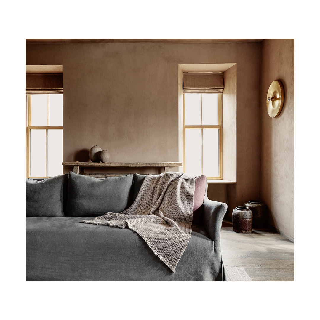 Interior of a living room featuring a gray sofa with throw blanket, neutral walls, natural light from windows, and minimal decorations.