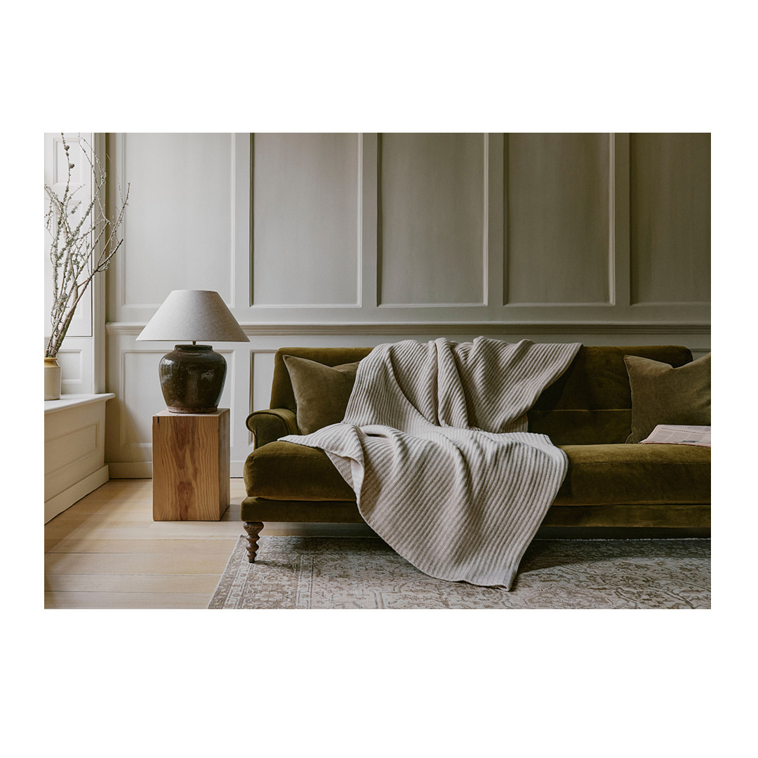 Interior shot featuring a green velvet sofa draped with a cream-colored ribbed blanket, accented by matching pillows and a wooden side table with a lamp.
