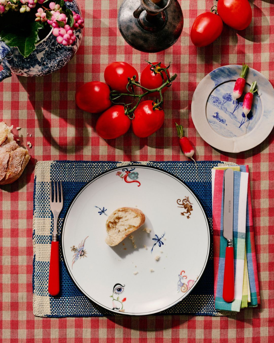 Overhead shot of a table setting with a plate of bread, fresh tomatoes on the vine, radishes, a utensil, and a floral centerpiece.