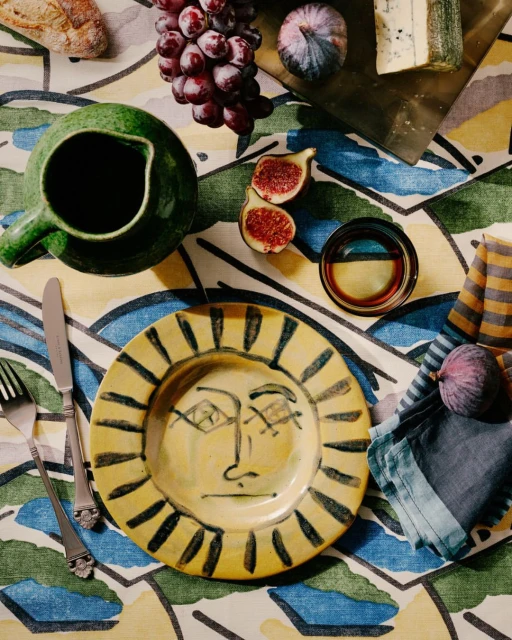 Overhead view of a yellow plate with a face design, alongside figs, grapes, cheese, and green pitcher on a patterned tablecloth.