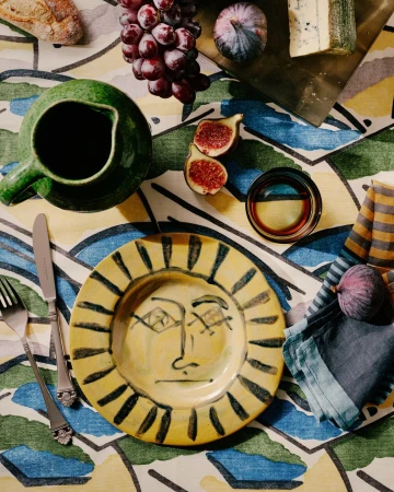 Overhead view of a yellow plate with a face design, alongside figs, grapes, cheese, and green pitcher on a patterned tablecloth.