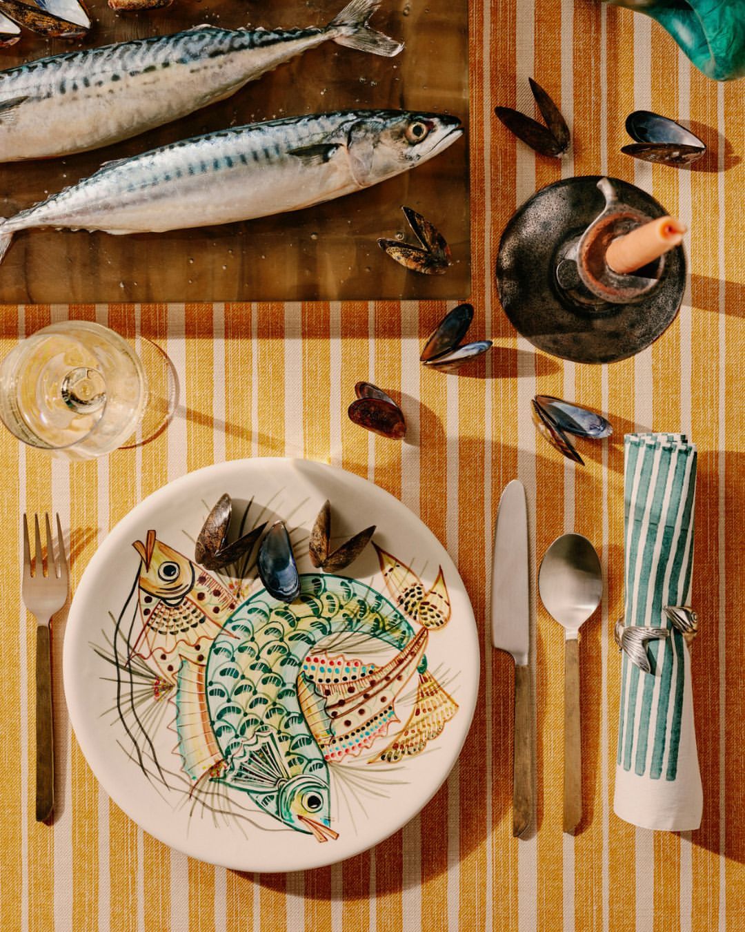Overhead shot of seafood dish with two mackerels, a ceramic plate with fish illustration, mussels, and silverware on striped fabric.