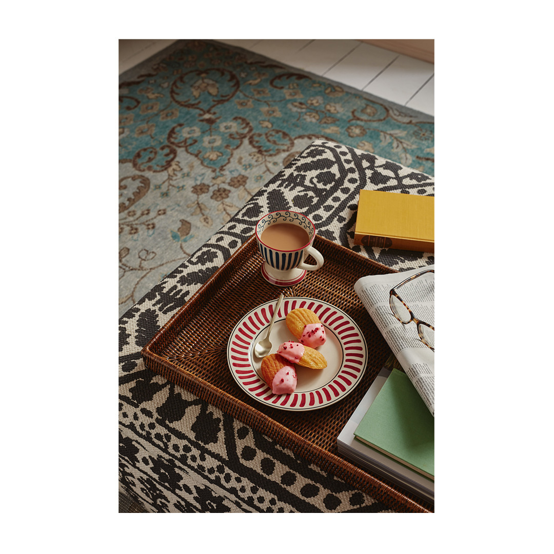 Tea and madeleines served on a wicker tray atop a patterned ottoman, alongside books and a throw, creating a cozy scene.