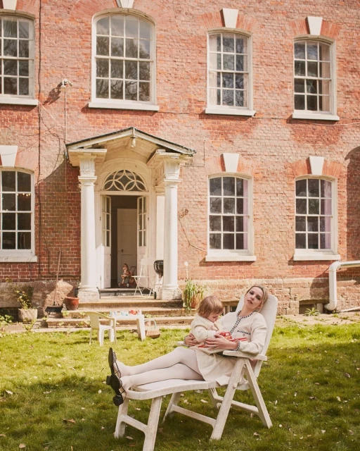 Woman and child relax on lawn chair in front of brick house; lawn and trees. Natural lighting, summer day, lifestyle image.