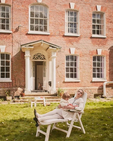 Woman and child relax on lawn chair in front of brick house; lawn and trees. Natural lighting, summer day, lifestyle image.