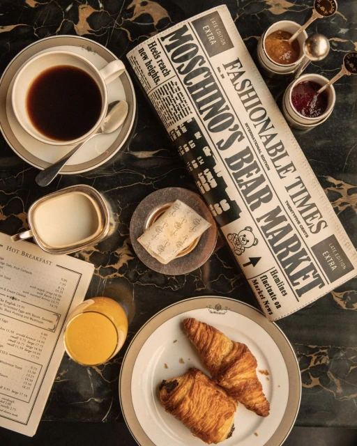 Overhead shot of coffee, juice, pastries, and a 'Moschino's Bear Market' newspaper on a marble table in a warm, inviting light.