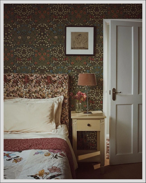 Floral-themed bedroom showcases bed with patterned headboard, nightstand with lamp and flowers, wallpaper, and a white door, creating cozy scene.