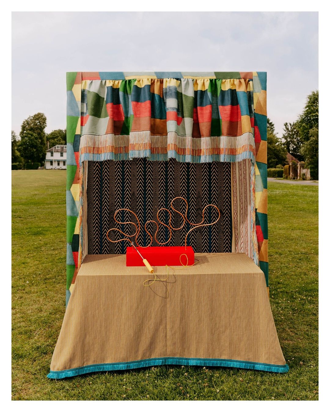 Handcrafted toy booth featuring a wire maze game on a draped table, set outdoors on a lawn in front of a house on a sunny day.