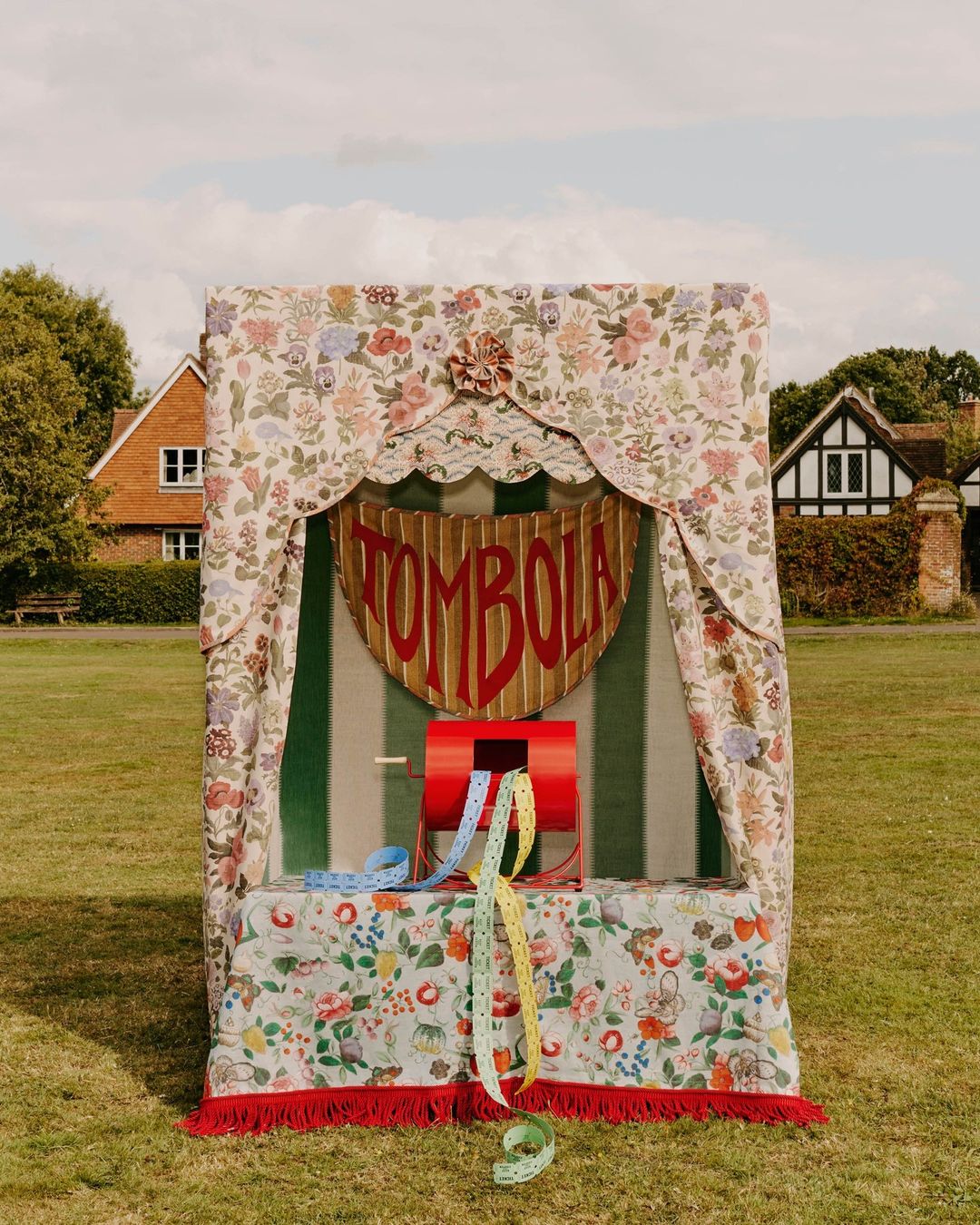 A vintage Tombola booth sits on a grassy lawn in front of houses, exuding classic charm and beckoning with the promise of chance and prizes.