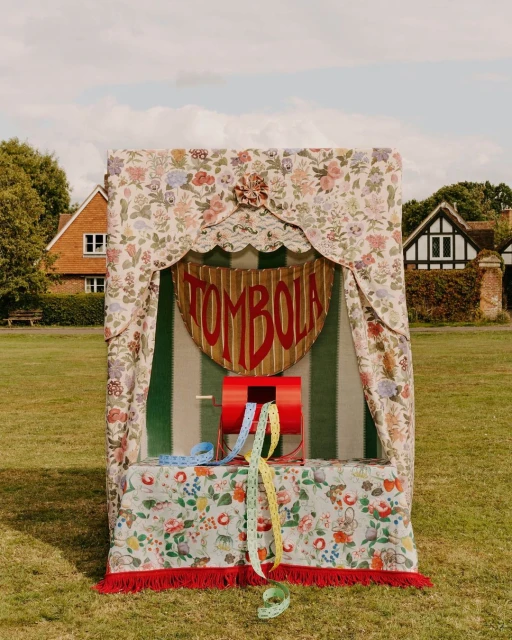 A vintage Tombola booth sits on a grassy lawn in front of houses, exuding classic charm and beckoning with the promise of chance and prizes.
