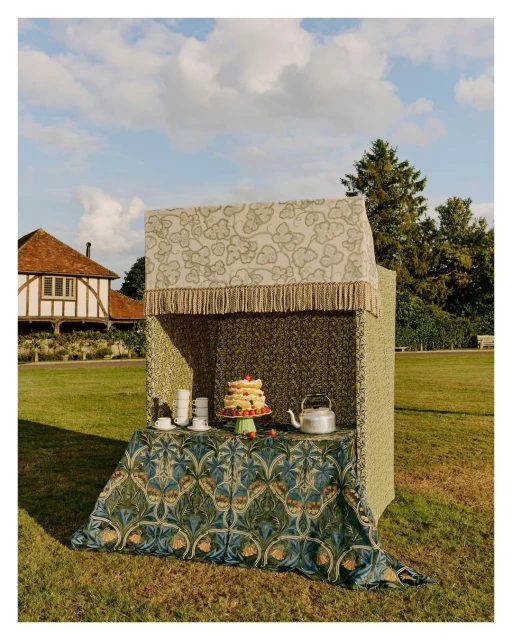 Outdoor display featuring a cake, teacups, and kettle on a patterned table, set against a green lawn and clear sky with a building behind.