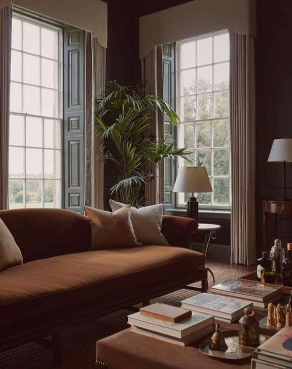 Cozy living room interior with a brown velvet sofa, decorative pillows, books on the coffee table, and natural light from large windows.