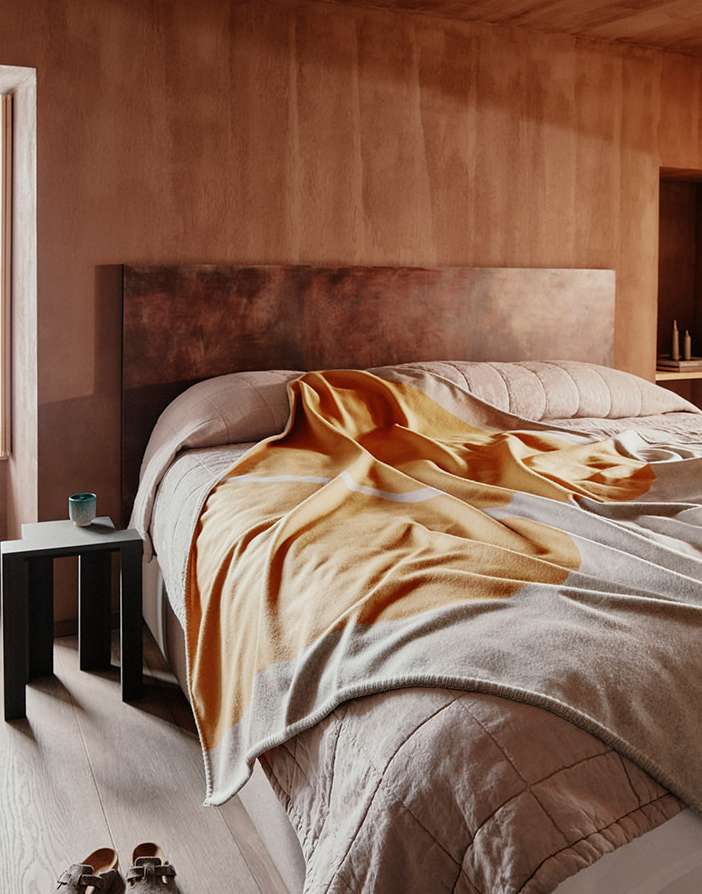 Interior bedroom with a bed covered in an orange and grey blanket, bedside table and wooden floor, and warm-toned plastered walls.