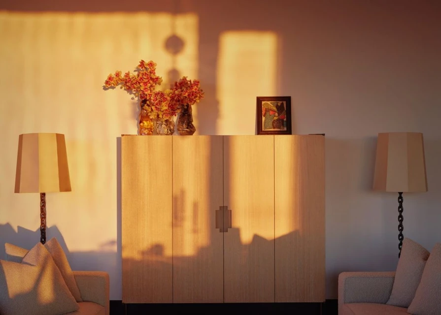 Interior room features a wooden cabinet, vases with red flowers, and lamps, creating a warm, inviting atmosphere with soft, golden light.