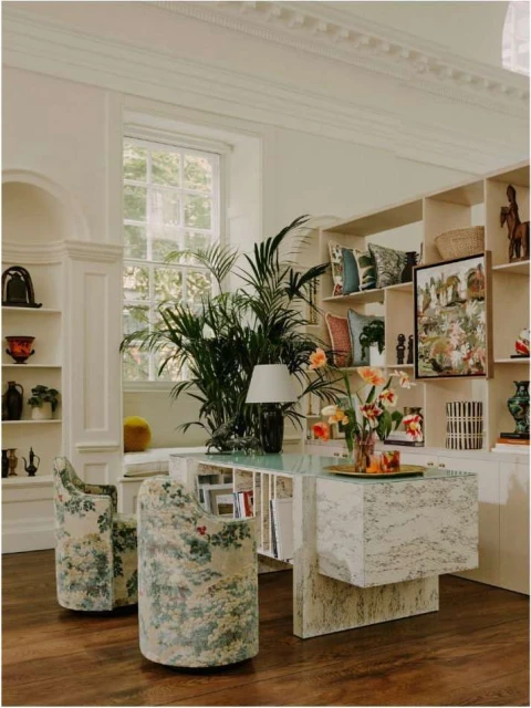 Home office interior featuring a marble desk, floral chairs, bookshelves, indoor plants, vases, and neutral wall colors. 