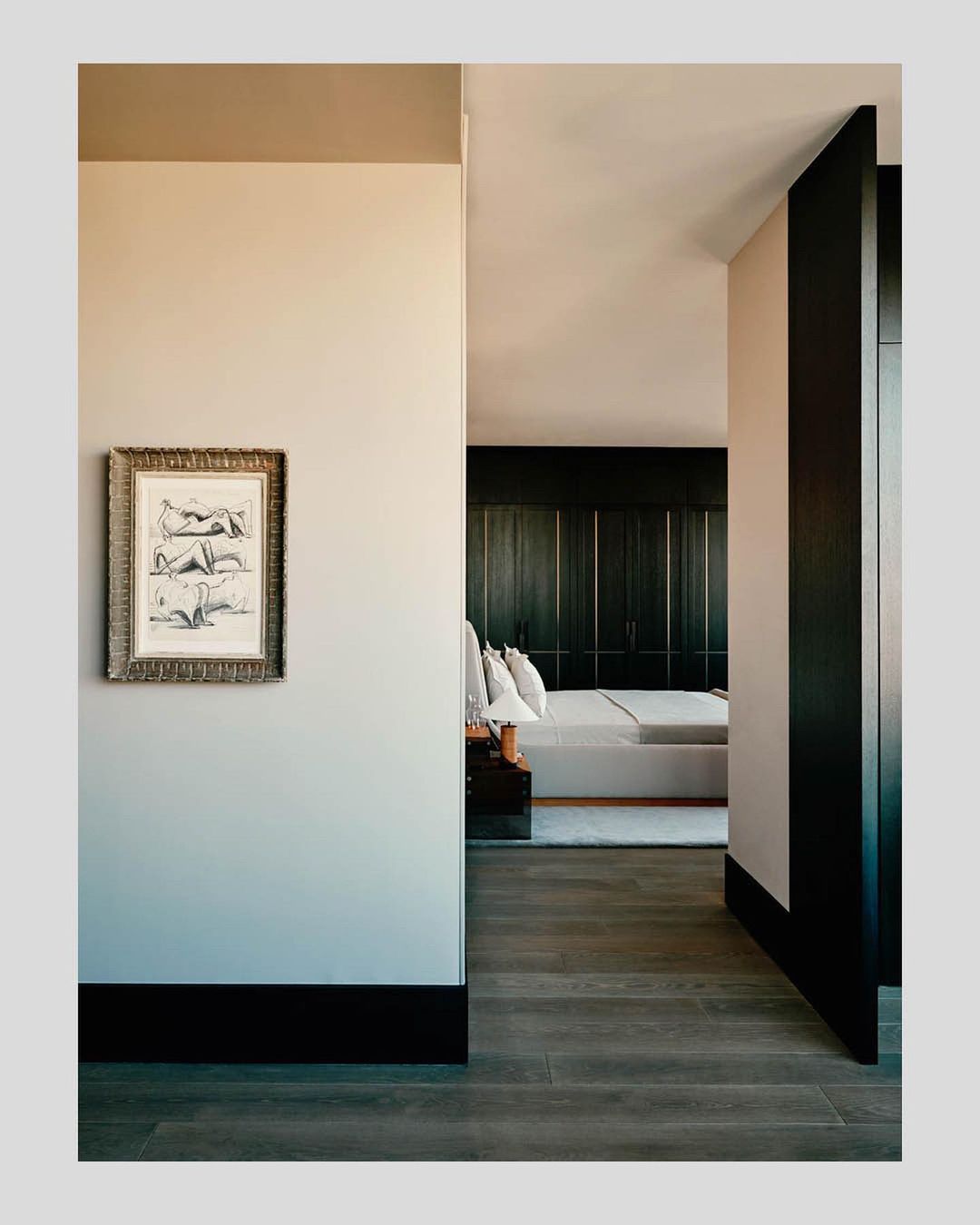 Hallway view into a modern bedroom with a framed artwork of reclining figures hanging on the wall, wood floors and trim details.