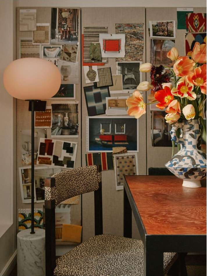 Interior design of a room with a lamp, flowers, table, and a chair with patterned upholstery against a wall with pinned images.